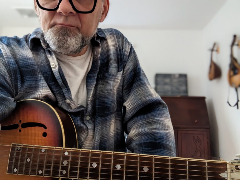 man with gray goatee wearing glasses sitting with guitar on his lap facing the camera in a workroom with mandolins on the white wall behind